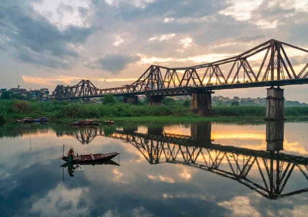 Cầu Long Biên lúc bình minh, in bóng xuống mặt sông với một chiếc thuyền nhỏ đi qua bên dưới